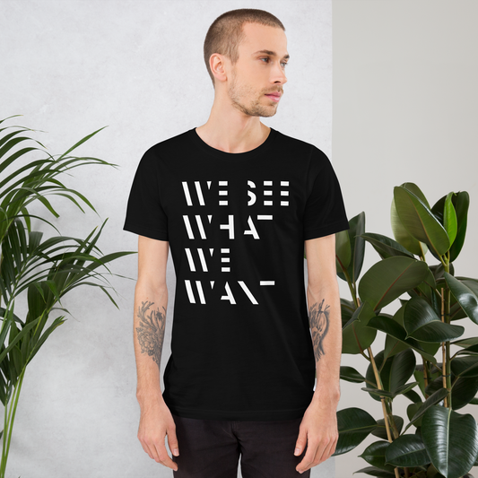 Mann trägt Schwarzes Unisex T-Shirt mit We See What We Want Print bei dem die Buchstaben teilweise ausgeschnitten sind